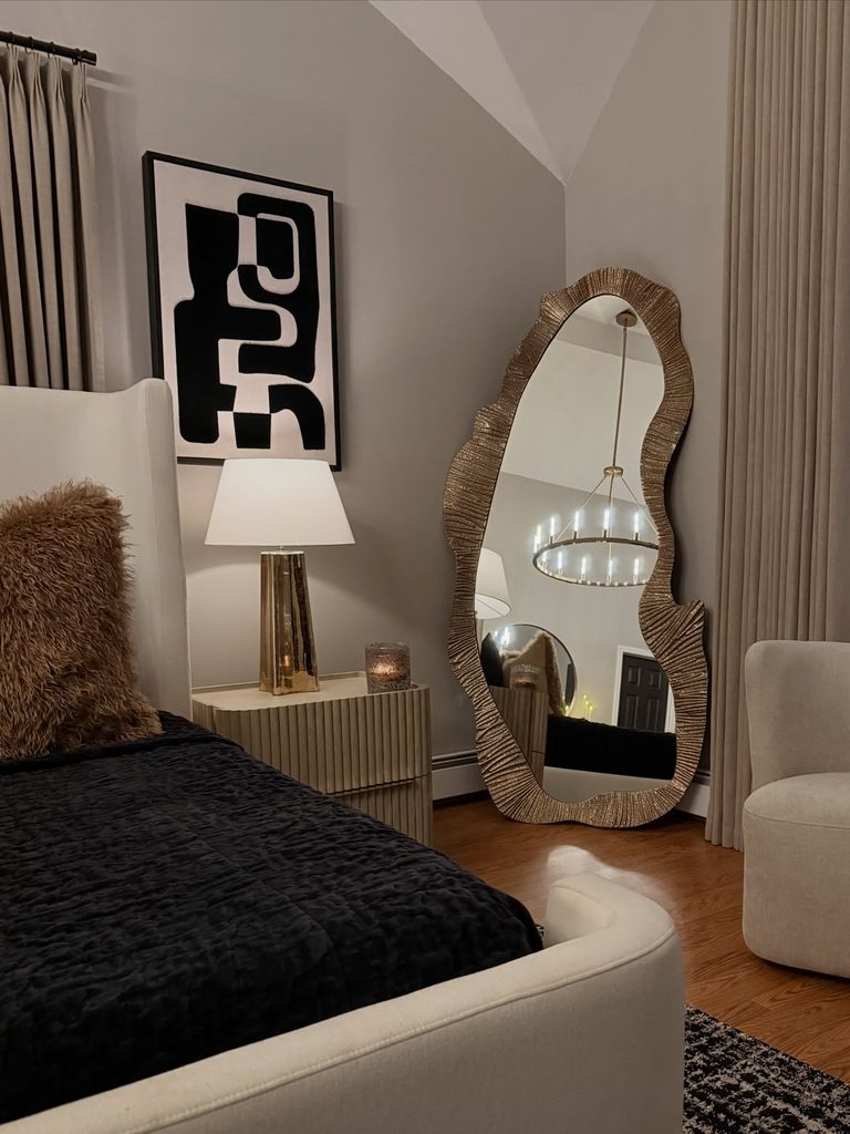 Bedroom with gold wavy floor mirror, white wingback bed, and black abstract wall art