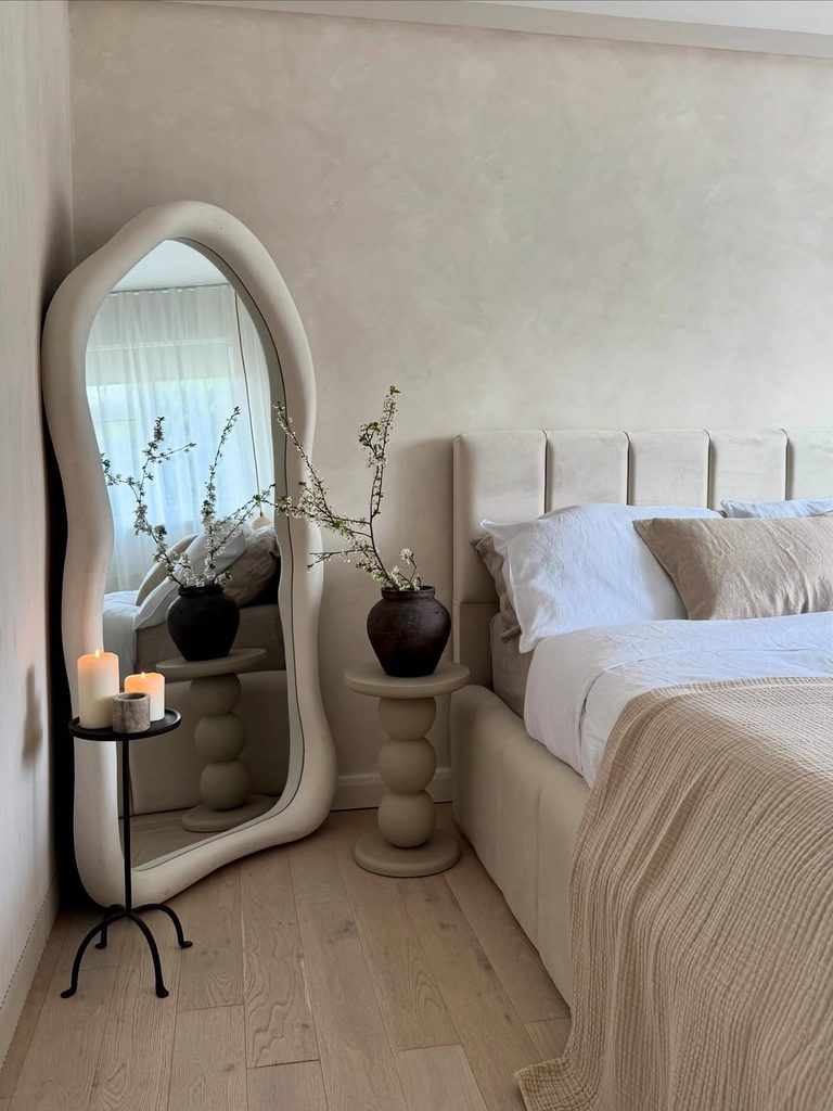 Neutral bedroom with arched floor mirror, bobbin nightstand, and dark ceramic vase on light oak floor