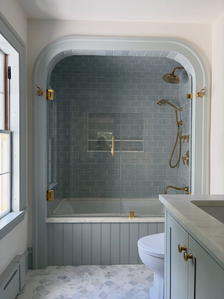 Blue gray subway tile shower alcove with unlacquered brass fixtures and glass enclosure