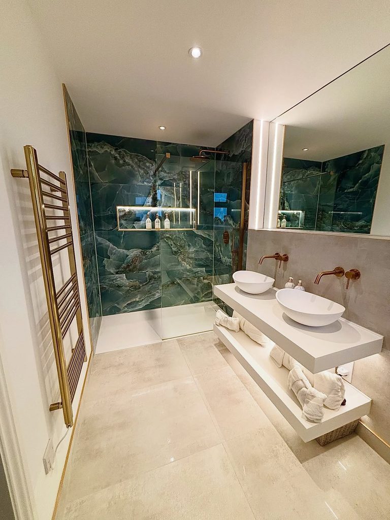 Luxury bathroom with teal green onyx shower tiles, double vessel sinks, and brushed brass towel rail